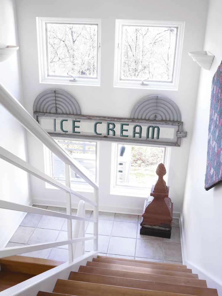 The wife used industrial materials when designing the house, including this stair railing. While not unheard of today, when she incorporated these elements in the early 1990s, it was avant-garde for residential homes. The solid-cherry open treads allow light through, augmenting the ethereal aesthetic. The metal Art Deco ice cream trade sign, offset by an architectural finial and hooked rug, was acquired from dealer Judy Milne.