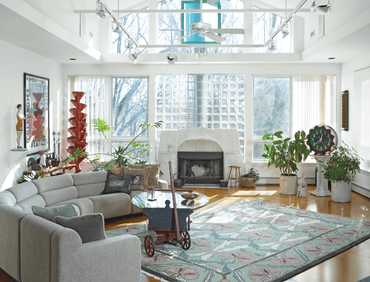 The peak of the living room ceiling shares the same profile as that of the atrium, which connects the front and back of the house visually. Glass bricks back the fireplace and add texture to the glass window wall. The fireplace surround, with hand-formed porcelain slabs and torn blocks of unglazed porcelain, is by contemporary artist/potter Paula Winokur, whose work is in the Philadelphia Museum of Art and elsewhere. A translucent Scalamandre silk screen, one of three in the room, creates the effect of stained glass. To the right is an early twentieth-century diner clock on a pedestal; opposite is a standing red sculpture that in reality is a painted metal grain chute that the couple purchased at the New York Pier Antique Show because of its visual impact. The rug has a William Morris style design, and the coffee table is a painted goat cart with modern glass top.