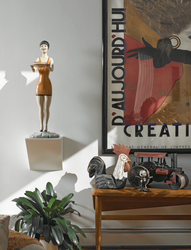 “This grouping of objects explains what we’re about,” says the wife. “We wouldn’t have had this sort of display in our previous house, but we now have the freedom to have fun in creating such vignettes.” Shown are a mid-century Danish Finn Juhl table, a circa-1940s carved bathing beauty figure, a papier-mâché rooster, and a Felix-the-Cat figure and a toy steam roller, set against a French poster.