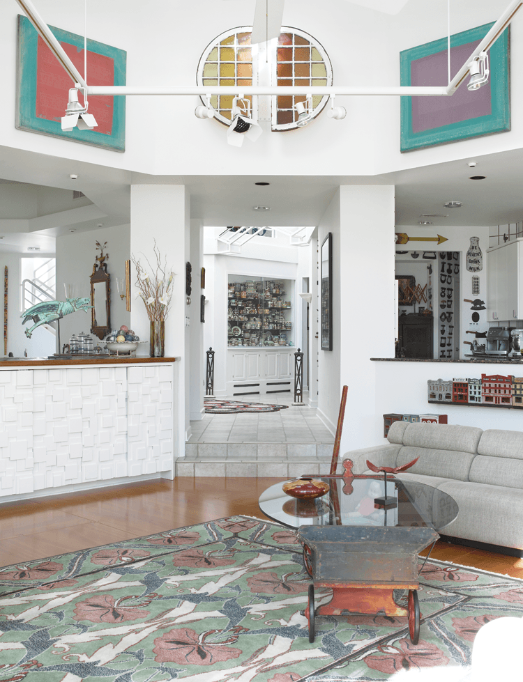 Looking toward the front of the house from the living room, two of the Scalamandre silk screens center two stained-glass panels. A wooden village from Wilsonboro, Pennsylvania, and made for a child’s toy train track, is mounted on the wall beneath the open kitchen. A portion of the mocha collection is visible through the corridor, and the dining room is on the left; a nineteenth-century French gargoyle downspout greets people as they walk into the living room.