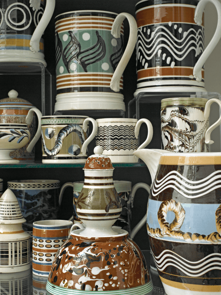 A dominant decorative technique shown here is the undulating “squiggle” designs created from slip trailing (a slurry of clay poured through multiple goose quills from a slip cup while the object rotates on a horizontal lathe). Another decorative device, present on the jug in foreground, is the coiled cable or earthworm pattern on a blue slip band. The unusual use of “off-set printing,” whereby an imprint is made from a ring dipped in slip, is evident on the mug in the upper left corner. A very early piece, circa 1770–1775, in the second row, far right, exhibits slip marbled and combed patterning; the “kick” at the base of the applied handle, seen on other English ceramics of the period, is extremely rare on mochaware. The collection includes two such examples.