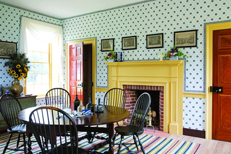 The Schorsches enjoy most meals in this bright and cheerful room enlivened by “Adena Pin Ring” pattern reproduction wallpaper, fixed dimity drapery, and striped “list” carpet. The gate-leg mahogany dining table is Philadelphia, circa 1720. Examples of archaeological material found on the site are displayed on the table for purposes of this photograph.