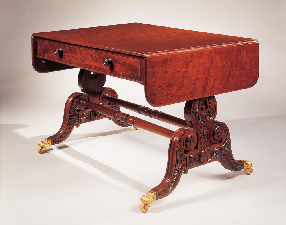 Classical carved mahogany sofa table with “plumb-pudding” mahogany top, Philadelphia, circa 1825. H. 28, W. 59© (leaves open), D. 30˙ in. Courtesy of Carswell Rush Berlin, Inc., New York. The beautiful countersunk foliate carving characteristic of Philadelphia, closely related to the carving on the previously illustrated sofa, can be seen in the down-swept legs and lyre base of this rich “plumb-pudding”-top sofa table. The double stretcher is also a typical device of Philadelphia table construction. The sofa table form, introduced by Thomas Sheraton in his 1803 Cabinet Dictionary, is extremely rare in American furniture. The profile of the scrolled legs were options in plate 5 of the Philadelphia Cabinet and Chair Maker’s Union Book of Prices for Manufacturing Cabinet Ware (Philadelphia, 1828).