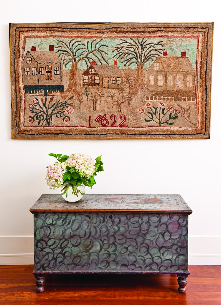 Within the master bedroom, a blue-painted Pennsylvania blanket chest, with whimsically enhanced black swirls, scallop, and dot decoration, sits beneath a large New England hooked rug, dated 1892.